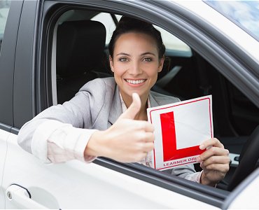 Young female driving lesson student with thumbs up sat in car holding a learner plate